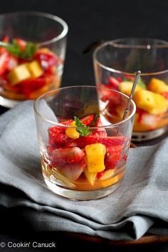 two glasses filled with fruit salad on top of a gray cloth next to silverware