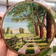 a person holding up a hand embroidered picture with trees and flowers in the foreground