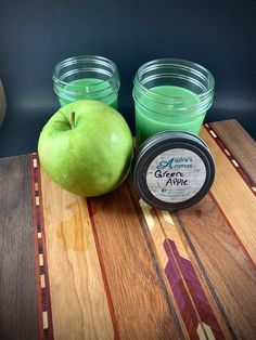 an apple sitting on top of a wooden cutting board next to two jars filled with green liquid