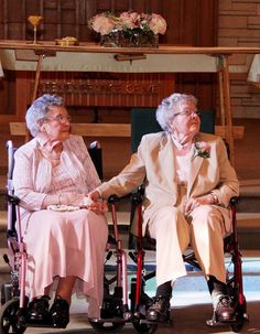 two elderly women sitting next to each other on wheelchairs in front of a wooden wall