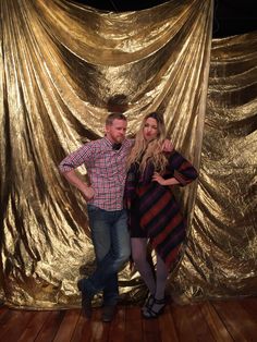 a man and woman standing in front of a gold curtain with their arms around each other