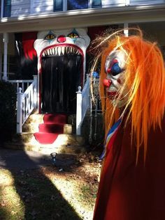 a person with orange hair and makeup standing in front of a house wearing a scary clown mask