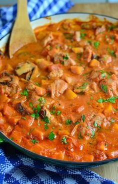 a pan filled with meat and vegetables on top of a blue table cloth next to a wooden spoon