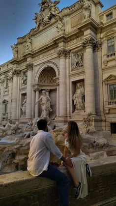 a man and woman sitting on a ledge in front of a building with statues around it