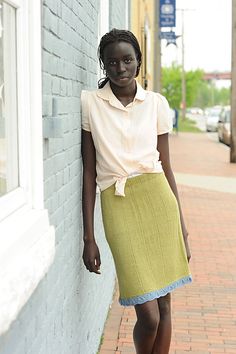 a woman leaning against a brick wall wearing a yellow skirt and white shirt with her hands in her pockets