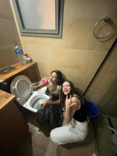 two women sitting on the floor in a bathroom next to a toilet and trash can