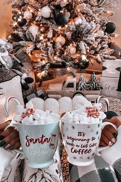 two coffee mugs with hot chocolate in front of a christmas tree and other decorations