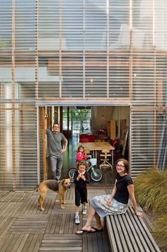 three people and a dog are sitting on a wooden bench in front of a house