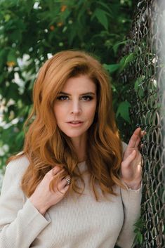 a woman with red hair leaning against a chain link fence and looking at the camera