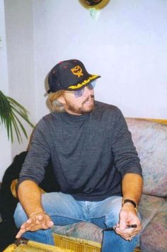 a man sitting on top of a couch holding a remote control in his hand and wearing a hat