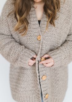 a woman standing in front of a white wall wearing a gray cardigan sweater and jeans