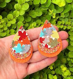 a hand holding two pairs of orange and gold earrings with leaves on them, in front of green foliage