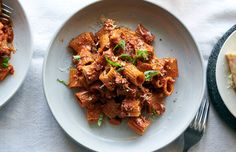 two white plates with pasta and meat on them next to a slice of cheese bread