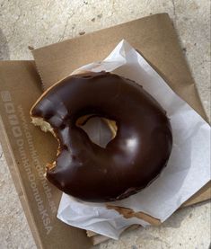 a half eaten chocolate donut sitting on top of a brown paper bag next to a cardboard box