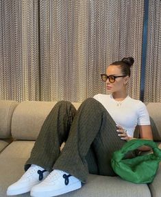 a woman sitting on top of a gray couch next to a green bag and white sneakers