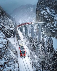 a red train traveling over a snow covered mountain next to a tall stone bridge in the middle of winter