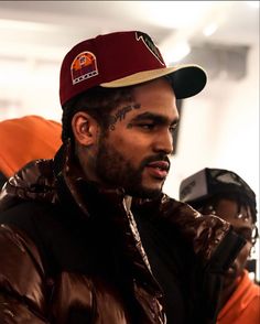 a man wearing a maroon hat and brown jacket with tattoos on his face looking off to the side