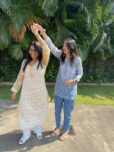 two women standing next to each other in front of some trees and bushes, one holding her hand up