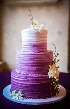 a three tiered purple cake with white flowers on the top and bottom, sitting on a blue plate