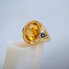 Antonini is a bold Modern era (circa 1990) citrine and tanzanite ring designed and created by famed Milanese jewelry house Antonini Jewelry. The 18k yellow gold ring centers a warm Oval Mixed Cut citrine gauged at approximately 8.50ct. The golden stone is flanked by two Pear Brilliant Cut tanzanites with the loveliest violet-blue hue. Antonini’s bezel set stones are framed by an open metal halo that follows the outline of the three-stone, lightening up the big & bold ring design. The ring is fin Bold Rings, Tanzanite Ring, Citrine Stone, 18k Yellow Gold Ring, Yellow Gold Ring, Blue Hues, Three Stone, Yellow Gold Rings, Bezel Setting