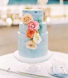 a three tiered blue cake with flowers on top and silverware next to it