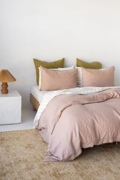 a bed with pink sheets and pillows in a white room next to a table lamp