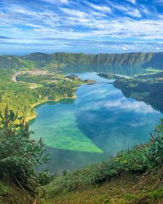 a lake surrounded by lush green trees and mountains under a blue sky with white clouds