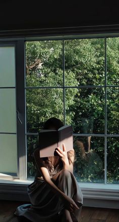 a woman sitting in front of a window holding a book up to her face while looking out the window