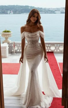 a woman in a white dress standing on a red carpet near the water and looking off to the side