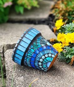 a blue and black stained glass vase sitting on top of a cement slab next to yellow flowers