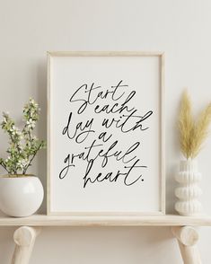 a white shelf topped with a vase filled with flowers next to a framed print that says stay calm and be grateful