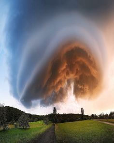 a very large cloud in the sky over a green field with trees and dirt road