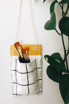 a white and yellow purse hanging on a wall next to a potted plant with two brushes in it