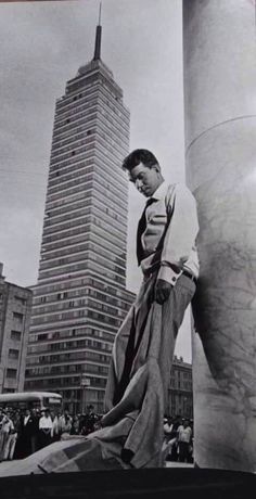 black and white photograph of a man leaning against a pillar in front of tall buildings