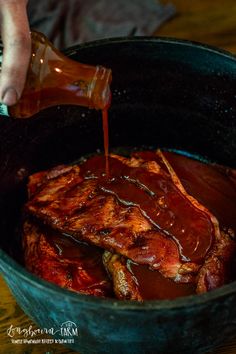 someone pouring barbecue sauce on top of ribs in a pot with other meats and vegetables