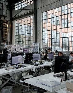 an office filled with lots of computers and people working on their desks in front of large windows