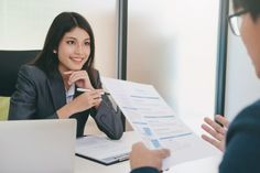 a woman sitting at a desk with a man in front of her talking to each other