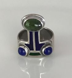 "There is a hint of arts and crafts, as well as, modernist in this unusual ring which you can wear with the Peridot facing up or down! The beautiful green stone, as well as the smaller vibrant blue Lapis are all cabochon and bezel set. The enamel is in wonderful condition. Aside from some slight wear, the ring is in overall fabulous shape. The band is almost 1/4\" wide, it is marked Sterling and has the initials CHM scratched on the silver behind the Peridot. Please see all photos for details, a Art Deco Green Emerald Multi-stone Ring, Modernist Rings With Inlay For Gift, Modernist Rings With Inlay Perfect For Gifts, Modernist Round Cabochon Jewelry, Modern Green Cabochon Jewelry, Unique Enamel Rings With Gemstone, Collectible Green Multi-stone Jewelry, Modernist Multi-stone Ring, Modern Multi-stone Green Rings