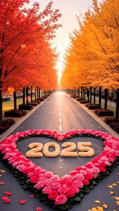 a heart shaped sign on the side of a road surrounded by flowers and trees in fall colors