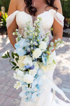 a woman in a wedding dress holding a bouquet of blue and white flowers with greenery