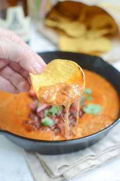 a hand holding a tortilla chip over a skillet filled with chili and cheese