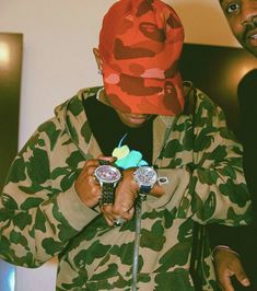 a man in camouflage jacket holding two watches and a red hat on top of his head