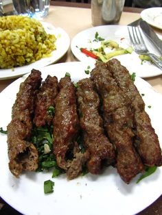 some meat is sitting on a white plate with greens and rice in the back ground