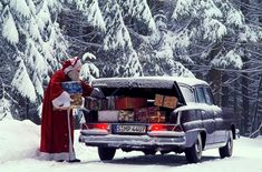 a man dressed as santa claus standing in the snow next to a car with presents on it