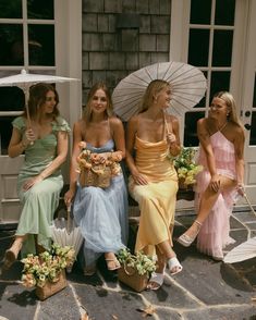 three women in dresses and umbrellas sitting on a stone patio with baskets of flowers