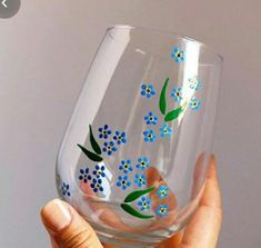 a hand holding up a wine glass with blue flowers painted on the side and green leaves