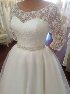 a woman wearing a wedding dress with sheer sleeves and beading on the waist, standing in front of a mirror