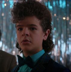 a young boy with curly hair wearing a suit and bow tie in front of a chandelier