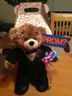 a brown teddy bear wearing a tuxedo holding a prom sign