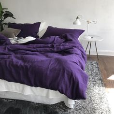 a bed with purple sheets and pillows in a bedroom next to a plant on the floor